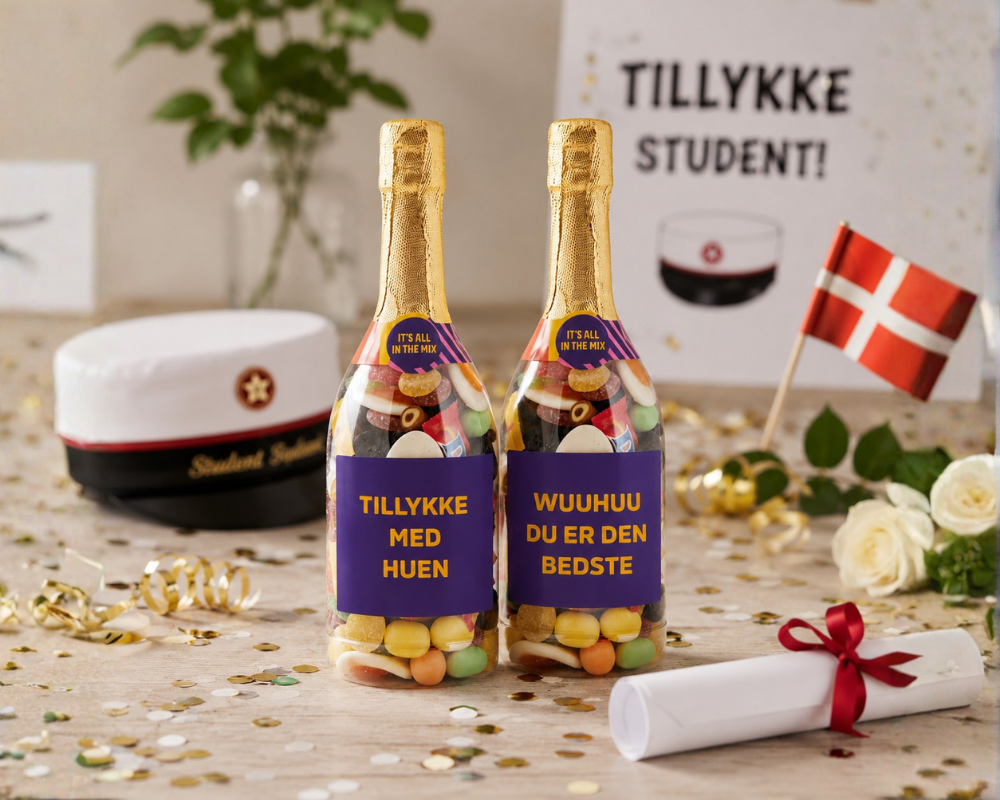 To champagneflasker fyldt med bland selv slik med studentertema omgivet af studenterhue, dansk flag, blomster og konfetti på festligt bord til fejring af studentertid.