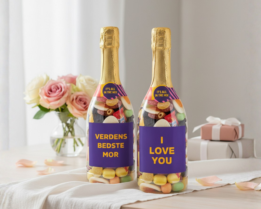 To champagneflasker fyldt med Bland Selv Slik fra SlikEkspressen med teksterne “Verdens bedste mor” og “I love you”, placeret på et elegant bord med blomster og gaveæsker i en lys og kærlig opsætning. Perfekt til mors dag.