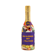 Champagneflaske fyldt med slik og lilla etiket med teksten "Du gjorde det – tillykke" – sød gave fra SlikEkspressen.